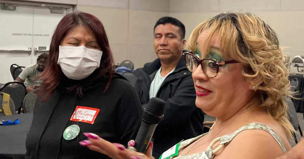 UE member Diana Martinez speaking into a microphone while a man and a woman behind her listen