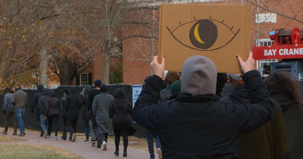 A single-file line of graduate workers seen from the back, the last one holding a cardboard sign with a giant eye drawn on it.