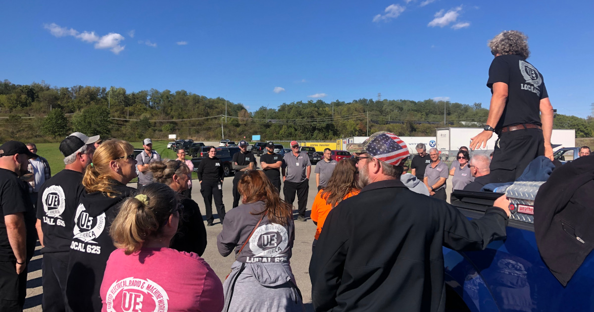 A large group of workers, many wearing UE t-shirts, gathered in a parking lot, listening to a speaker on the back of a truck.